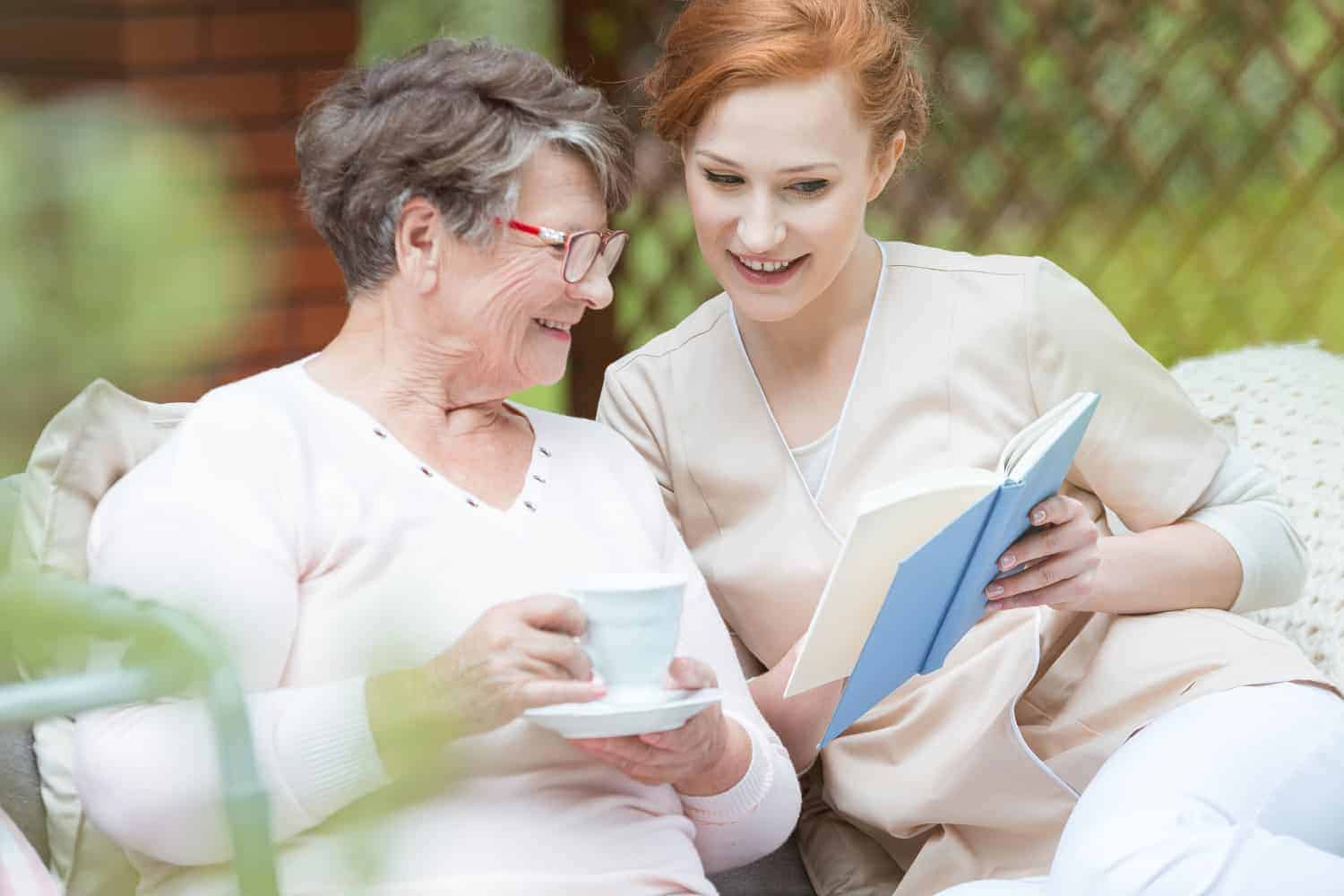 Generations sharing a moment: an elderly woman and a younger woman enjoying a warm conversation over a book and tea in a cozy garden setting, deepening their companionship.