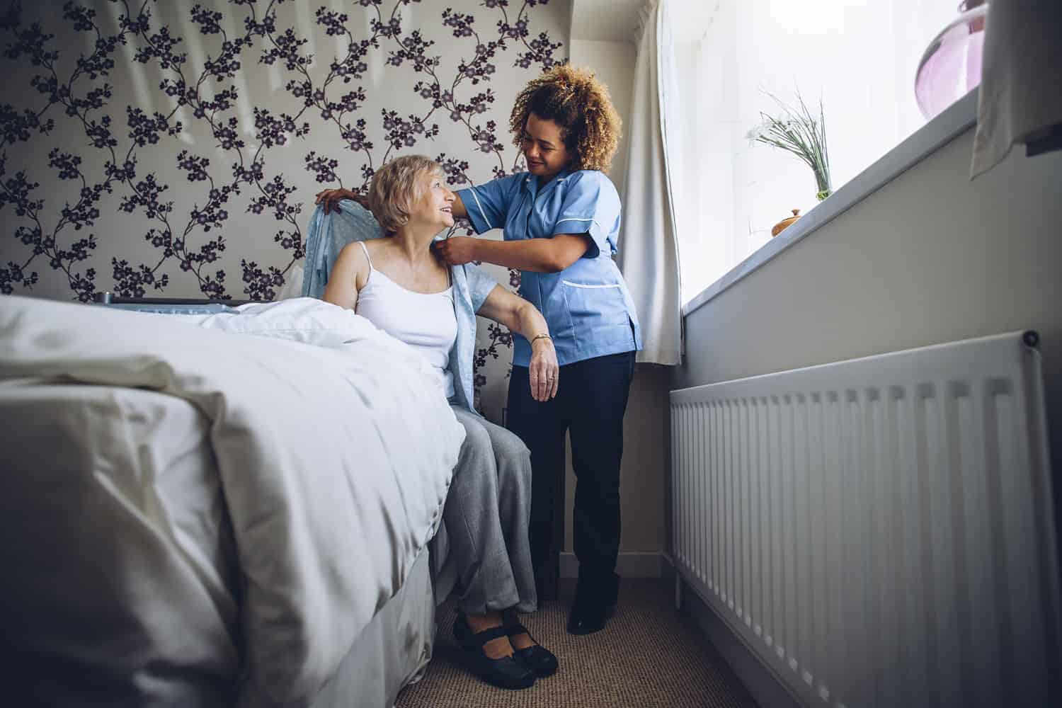 A compassionate homecare worker gently assists an elderly patient seated at the edge of a bed in a cozy, wallpapered room in Winkfield, Berkshire.