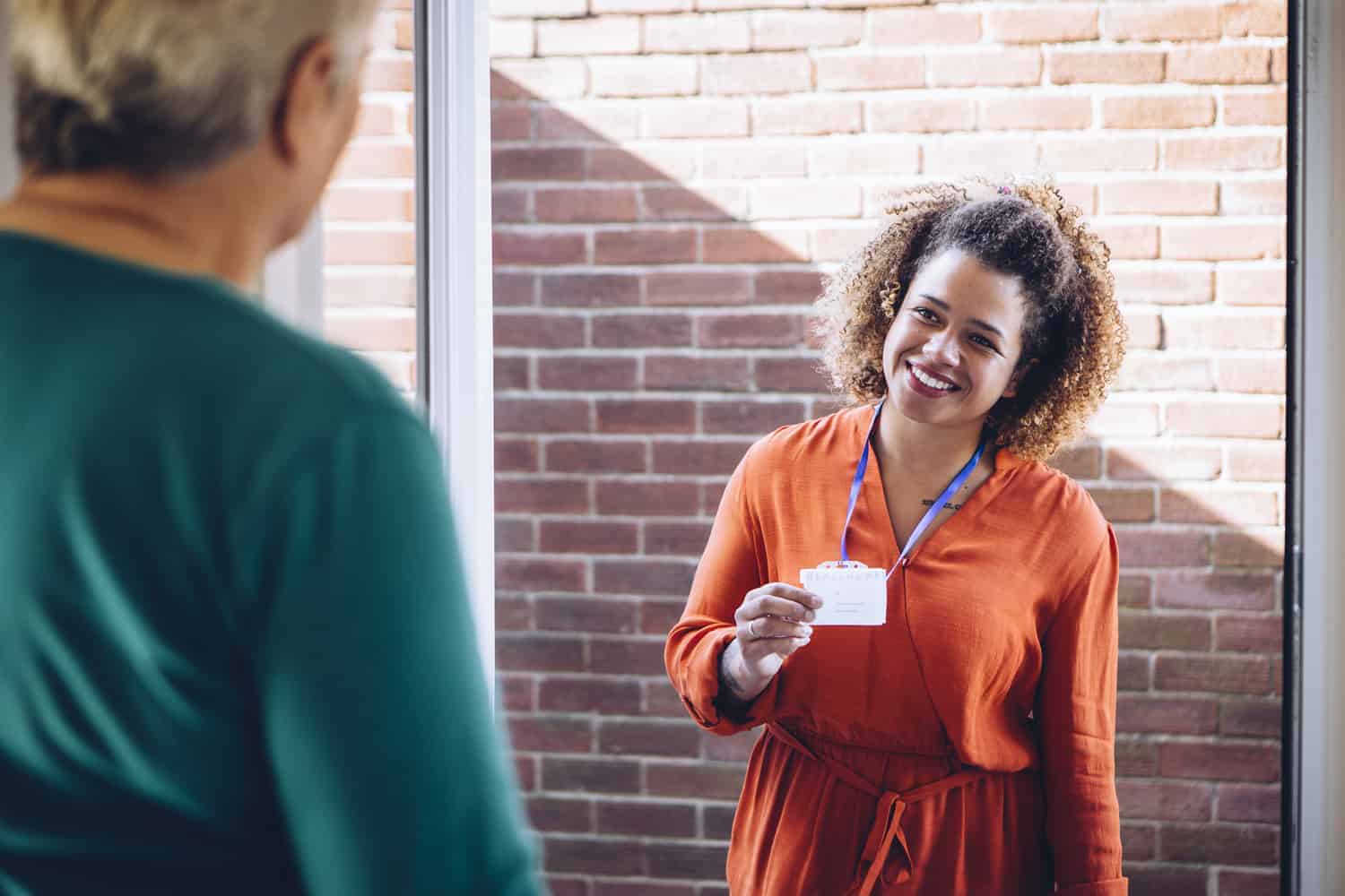 A smiling professional woman displaying her ID badge to a colleague at the entrance of a brick-walled office, ready to discuss tailored care programs.