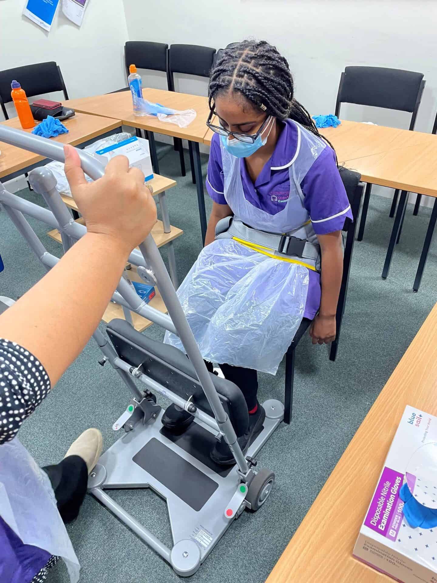 A healthcare student in a purple uniform practices transferring skills using a mechanical lift in a simulation lab, under supervision.