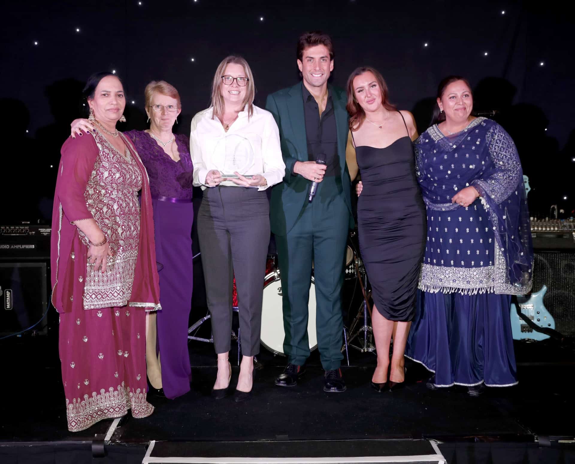 A group of five women and one man stand proudly on stage, smiling and holding a Regional Award from the Great British Care Awards. Dressed in formal, traditional attire, they pose against a starry backdrop with musical instruments behind them.