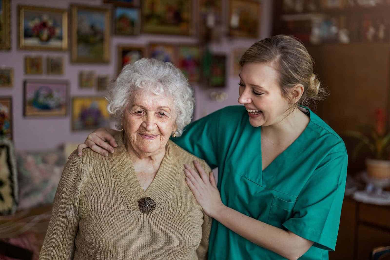 A smiling elderly woman with white hair and a beige sweater stands beside a younger person in a green medical uniform. In the room adorned with paintings, the young caregiver offers comfort, aware of the tender moments at end-of-life in dementia as they gently touch her shoulder.