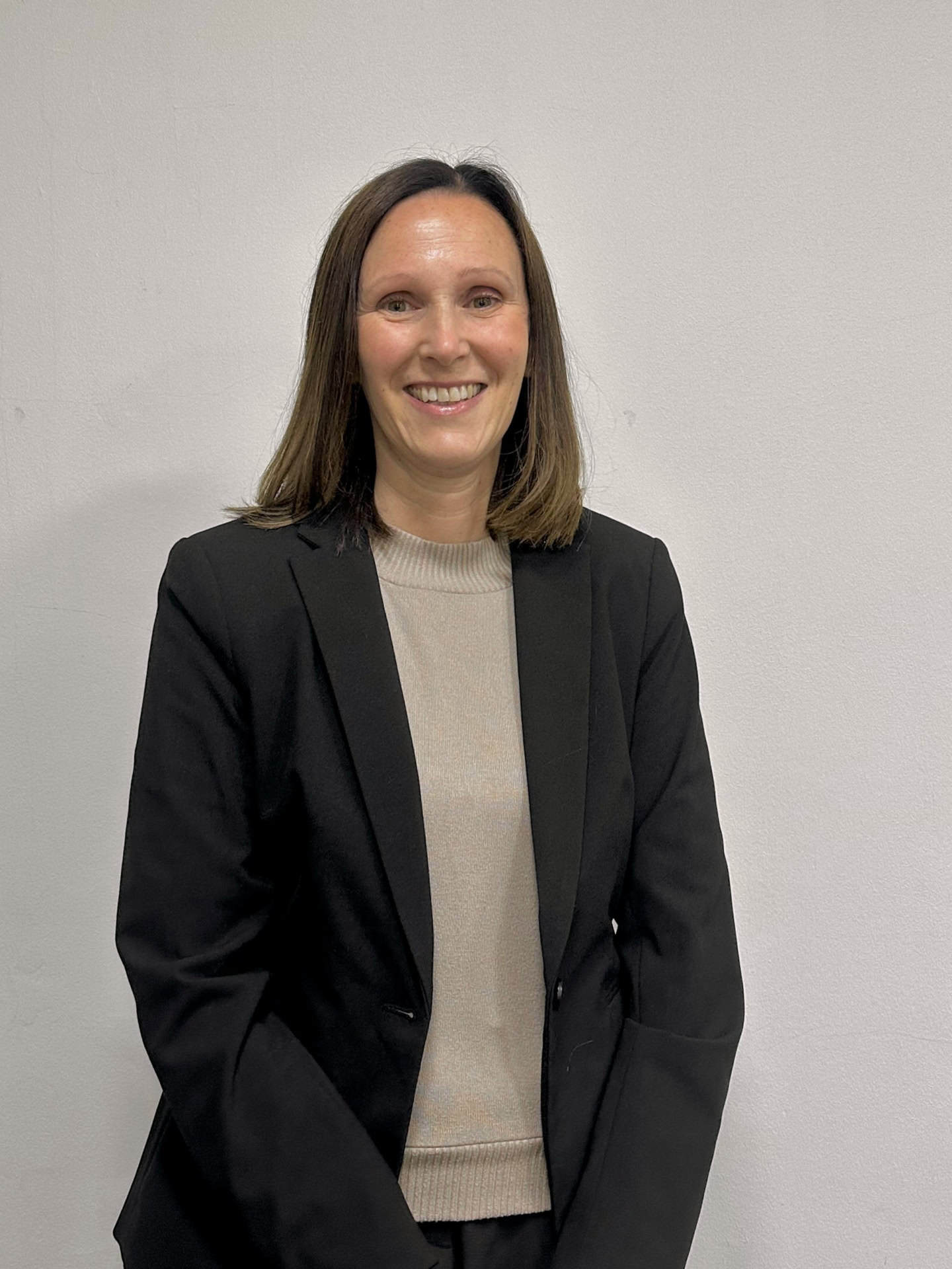 Meet our team: A woman with straight brown hair, wearing a black blazer over a light beige top, smiles while standing in front of a plain white wall.