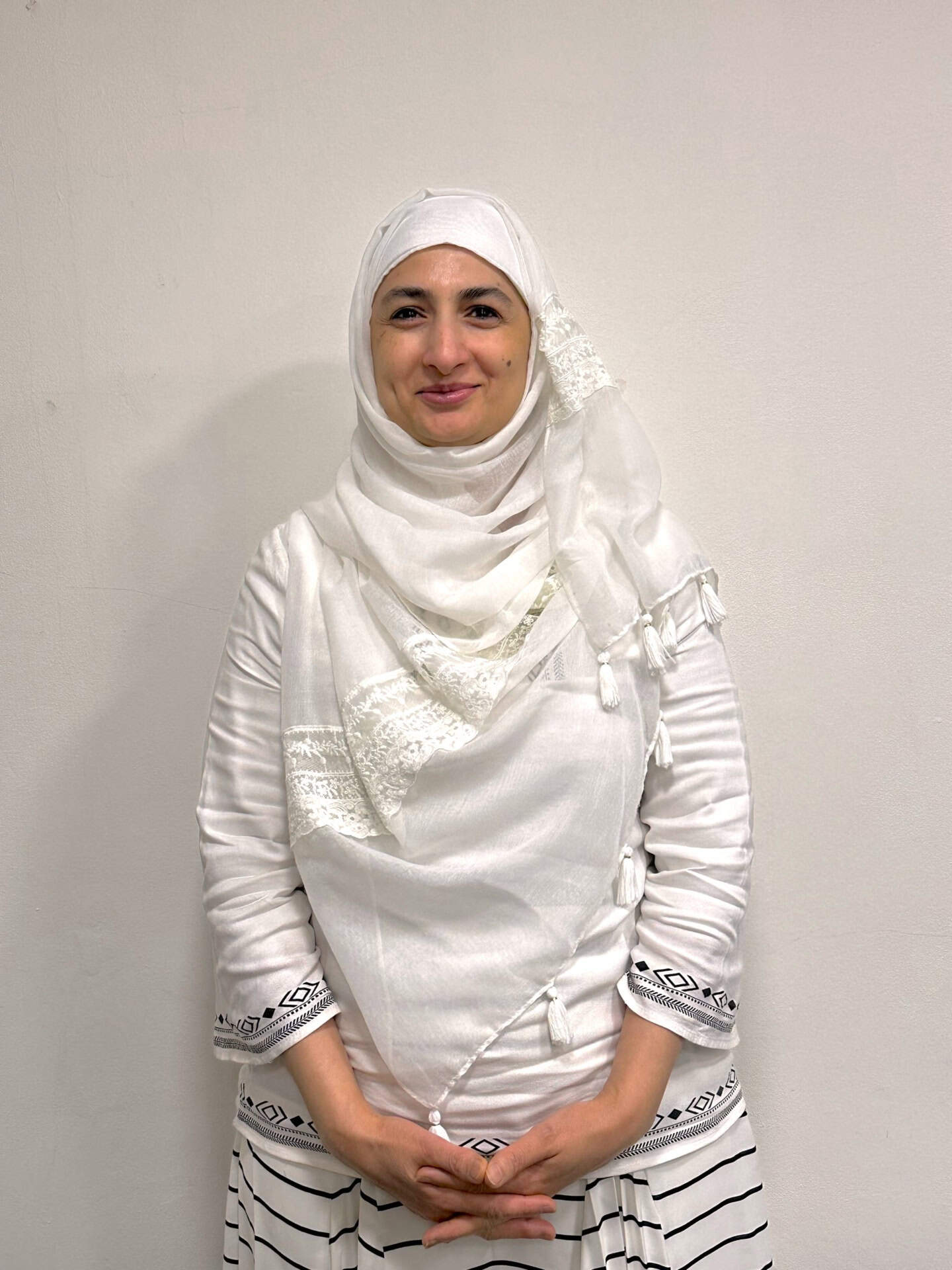 Meet our team: A woman wearing a white hijab and a white dress with black patterns on the sleeves and skirt stands against a plain light-colored wall, smiling gently with her hands clasped in front of her.