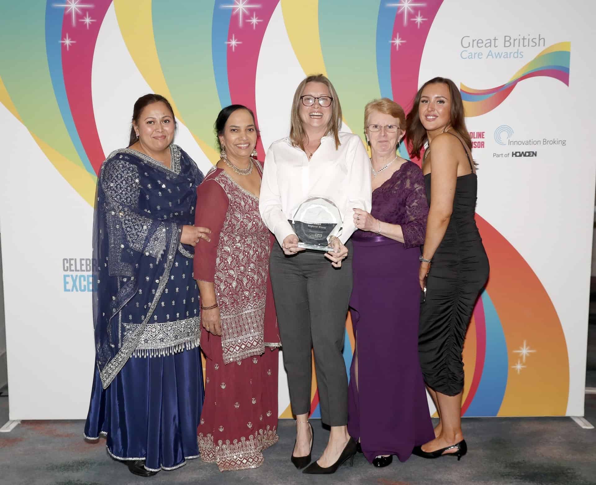 Five women stand together, smiling, with one holding a glass award. They are dressed in formal attire and are posing in front of a colorful Great British Care Awards backdrop.