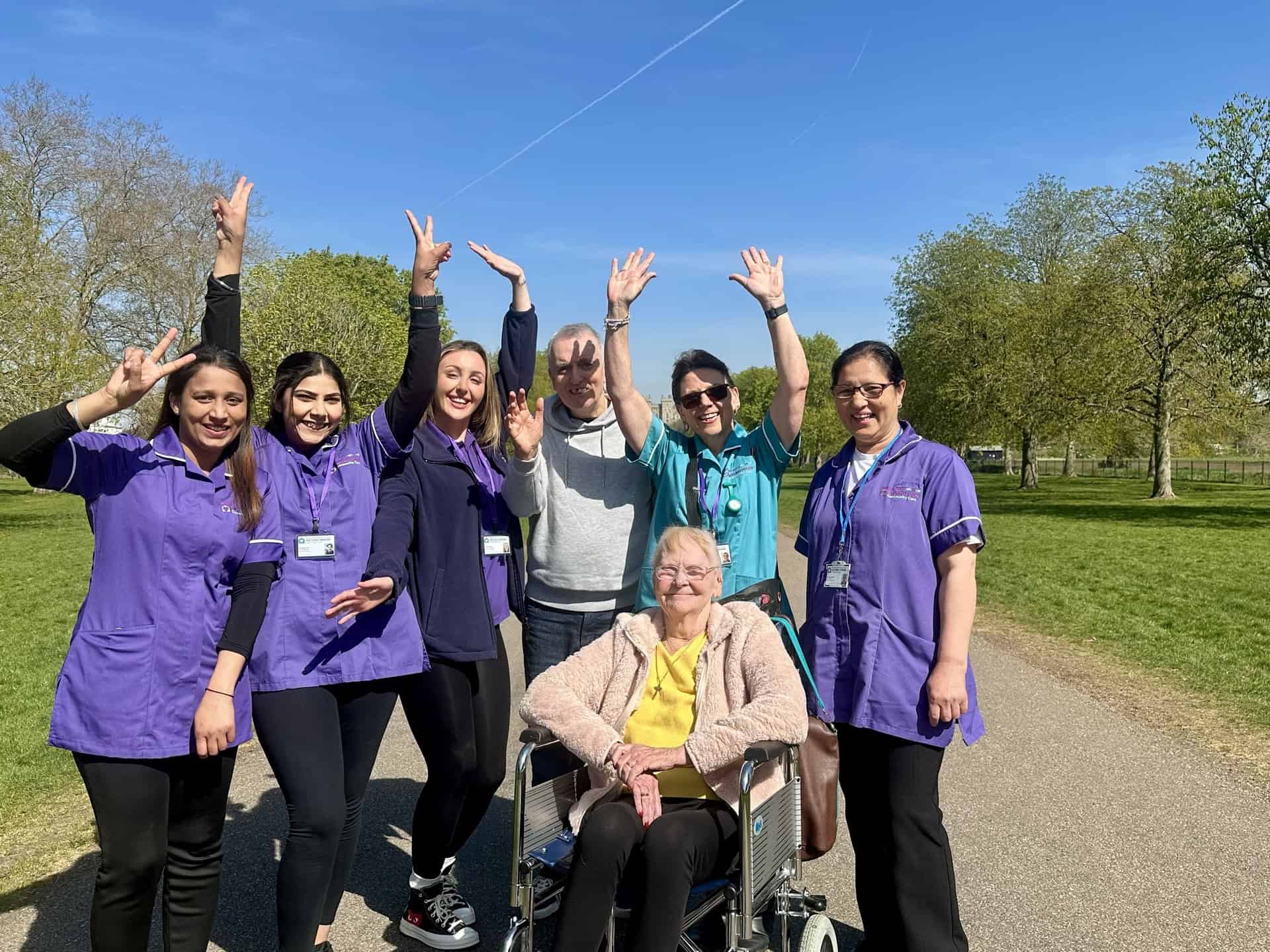 A group of smiling caregivers and an elderly woman in a wheelchair pose together outdoors on a sunny day, raising their hands and making peace signs in a park with green trees and blue sky.
