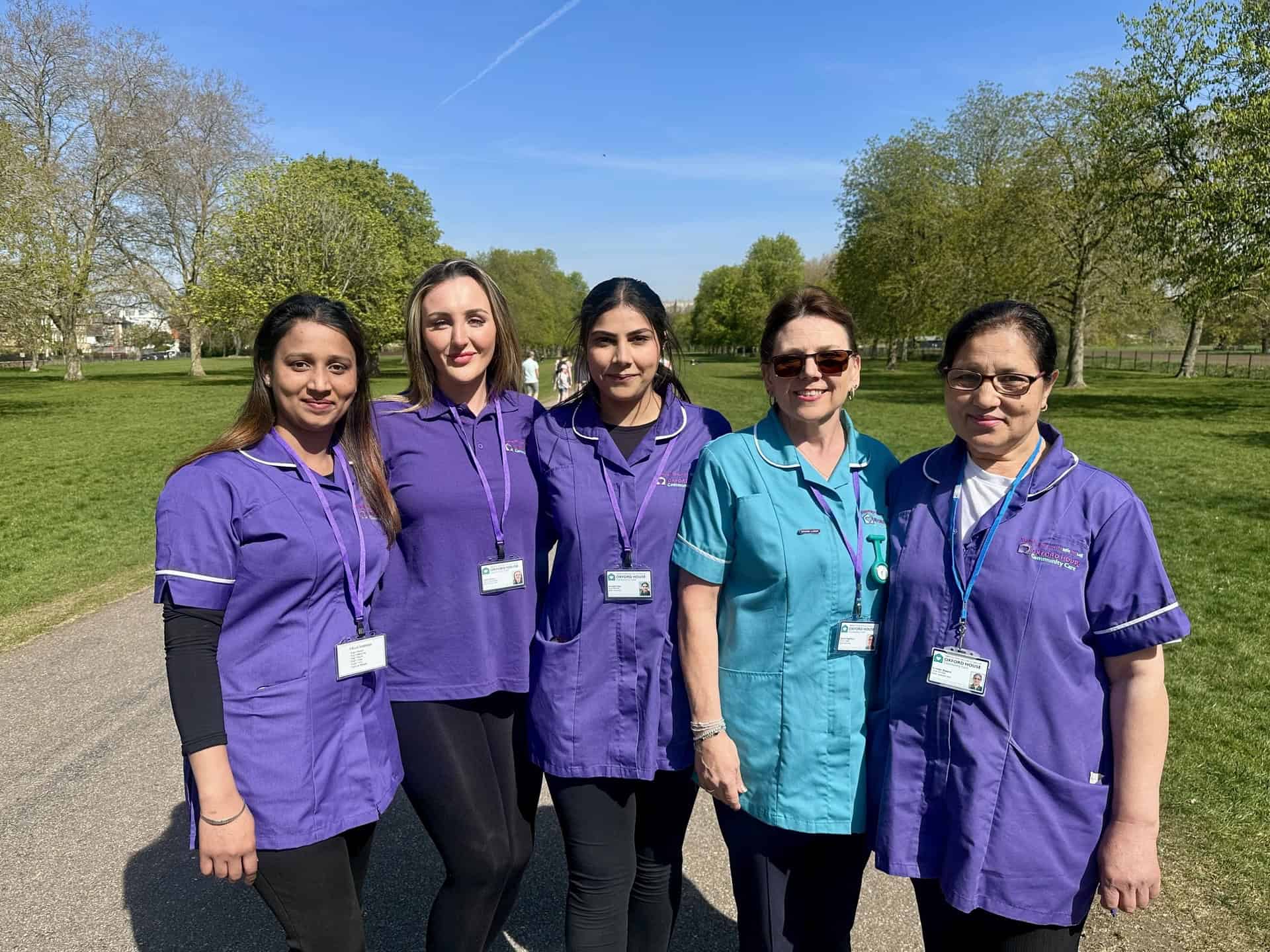 Five women, four in purple uniforms and one in a teal uniform, stand together and smile outdoors on a sunny day with green grass and trees in the background. They all wear name badges.