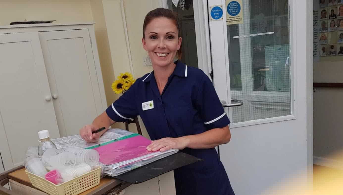 A smiling healthcare professional in blue scrubs standing at a counter while writing on a clipboard, with medical supplies nearby and a "Contact Us" poster board in the background.